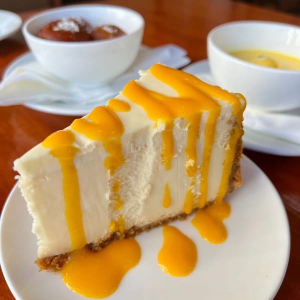 A slice of cheesecake with mango drizzle, alongside bowls of gulab jamun and mango lassi.