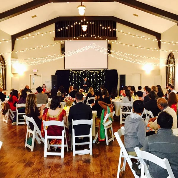 Guests seated at tables in a large hall with string lights and a speaker on stage.