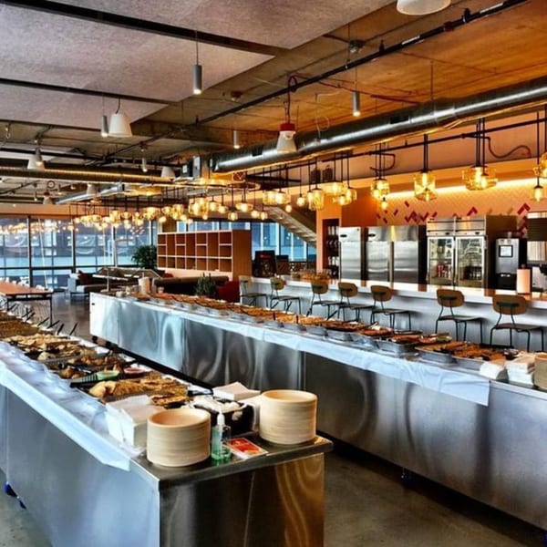 A modern cafeteria with long buffet lines filled with food, warm lighting, and large windows.