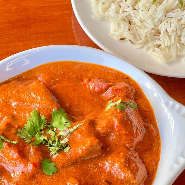 A white bowl of orange chicken tikka masala with cilantro, next to a plate of white rice.