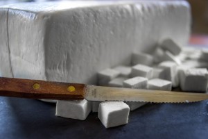 A block of fresh Paneer behind a bunch of tiny paneer cubes cut up with a knife.